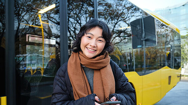 Person mit dunklen Haaren, dunkelblauer Jacke und braunem Schal steht vor einem gelben Bus mit reflektierenden Fenstern.