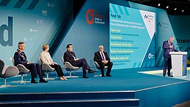 Drei Männer und eine Dame sitzen bei einer Podiumsdiskussion auf der Bühne. Der Moderator steht rechts am Rednerpult.