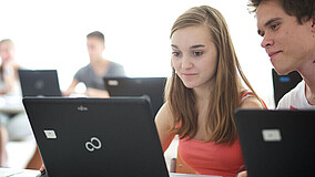 Eine junge Frau und ein junger Mann sitzen in einem Klassenzimmer nebeneinander, vor ihnen steht jeweils ein Laptop auf dem Tisch. Im Hintergrund sind weitere Personen und Laptops zu erkennen.
