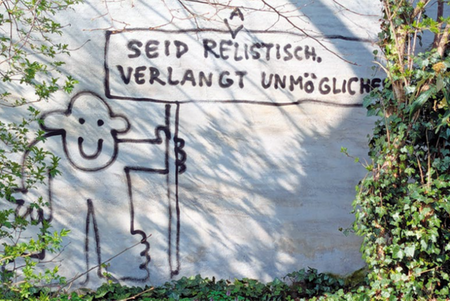 Graffiti an einer Wand zeigt eine stilisierte Figur mit einem Schild, auf dem steht 'SEID REALISTISCH VERLANGT UNMÖGLICHES'