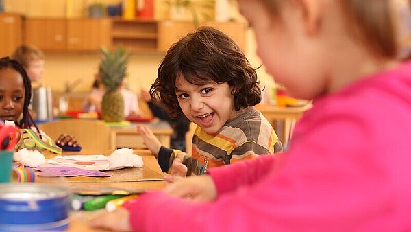 Kinder sitzen in einer IB-Kita an Tischen, während ein Junge in die Kamera lächelt.