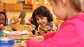 Kinder sitzen in einer IB-Kita an Tischen, während ein Junge in die Kamera lächelt.