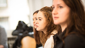 Drei junge Frauen sitzen nebeneinander in einem Klassenzimmer. Im Hintegrund sind verschwommen weiße Wände, ein Stuhl und eine weitere Person am linken Bildrand erkennbar.