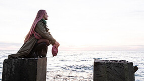Eine junge Frau hockt auf einem Steinblock, im Hintergrund ist das Meer zu sehen. Die Frau hat rosafarbene Haare und hält eine rosafarbene Tasche in den Händen.