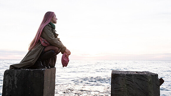 Eine junge Frau hockt auf einem Steinblock, im Hintergrund ist das Meer zu sehen. Die Frau hat rosafarbene Haare und hält eine rosafarbene Tasche in den Händen.