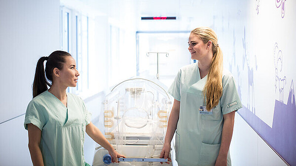 Zwei Frauen in medizinischer Arbeitskleidung schieben gemeinsam ein Babybett durch einen hellen Krankenhausflur