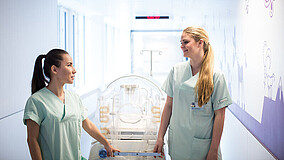Zwei Frauen in medizinischer Arbeitskleidung schieben gemeinsam ein Babybett durch einen hellen Krankenhausflur