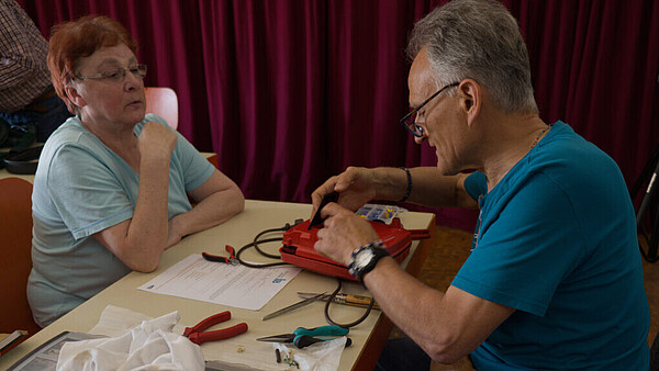Im Repair-Café des IB sitzt eine Frau neben einem Mann, der ein Gerät repariert.