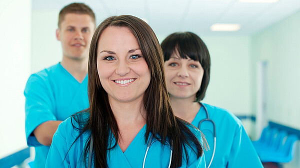 Drei Personen in blauen medizinischen Kitteln, vorne eine Frau mit Stethoskop, im Hintergrund zwei Personen, eine mit verschränkten Armen.