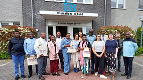 15 Personen mit schwarzer und weißer Hautfarbe stehen vor dem Haupteingang der Zentralen Geschäftsführung des IB. Der Eingang besteht aus einer weißgerahmten Glastür, beschriftet mit "Internationaler Bund" und "Valentin-Senger-Straße 5". Über der Tür befindet sich ein blaues IB-Logo.