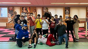 Eine Gruppe Jugendlicher und ein Mann in einer Turnhalle. Sie halten die Fäuste hoch und blicken in die Kamera.