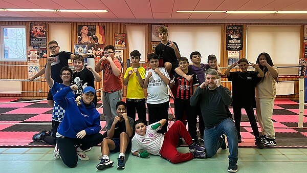 Eine Gruppe Jugendlicher und ein Mann in einer Turnhalle. Sie halten die Fäuste hoch und blicken in die Kamera.