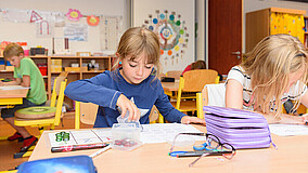Kinder sitzen im Klassenraum an ihren Tischen und schreiben oder basteln.