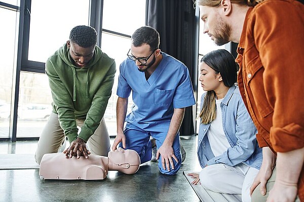 Zwei junge Männer knien auf dem Boden. Einer der beiden übt an einem Trainigstorso eine Herzdruckmassage. Daneben sitzen zwei weitere Personen auf dem Boden und schauen zu.