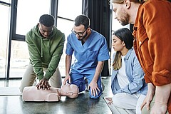Zwei junge Männer knien auf dem Boden. Einer der beiden übt an einem Trainigstorso eine Herzdruckmassage. Daneben sitzen zwei weitere Personen auf dem Boden und schauen zu.