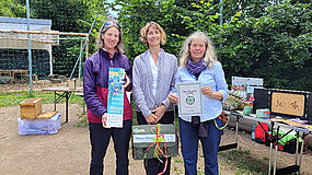 Drei Frauen stehen nebeneinander. Sie halten eine Urkunde und Geschenke in den Händen. Im Hintergrund sind Tische und Bäume zu sehen. 