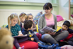 Fünf kleine Kinder sitzen zusammen mit einer jungen Frau auf Kissen auf dem Boden. Die Frau hält ein Buch in der Hand.