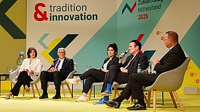 Fünf Personen sitzen auf Stühlen auf einer Bühne mit gelbem Boden vor einer Wand mit den Schriftzügen 'tradition & innovation' und 'Zukunftstag Mittelstand 2026.