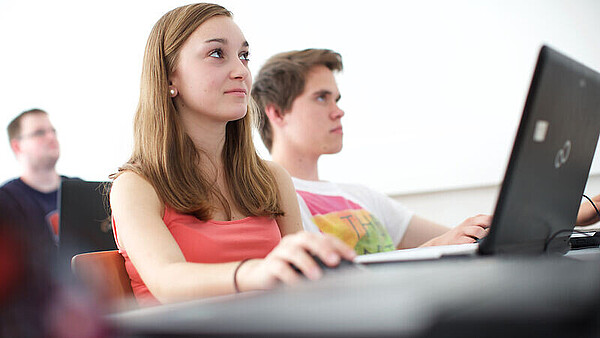 Eine Schülerin und ein Schüler sitzen nebeneinander im Klassenzimmer. Im Hintergrund sitzt ein weiterer Schüler. Vor der Schülerin, die sich im Vordergrund befindet, steht ein aufgeklappter Laptop.