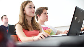 Eine Schülerin und ein Schüler sitzen nebeneinander im Klassenzimmer. Im Hintergrund sitzt ein weiterer Schüler. Vor der Schülerin, die sich im Vordergrund befindet, steht ein aufgeklappter Laptop.