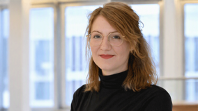 Jüngere Frau mit längeren Haaren und einer Brille. Sie trägt einen schwarzen Pullover. Im Hintergrund ist eine Fensterfront.