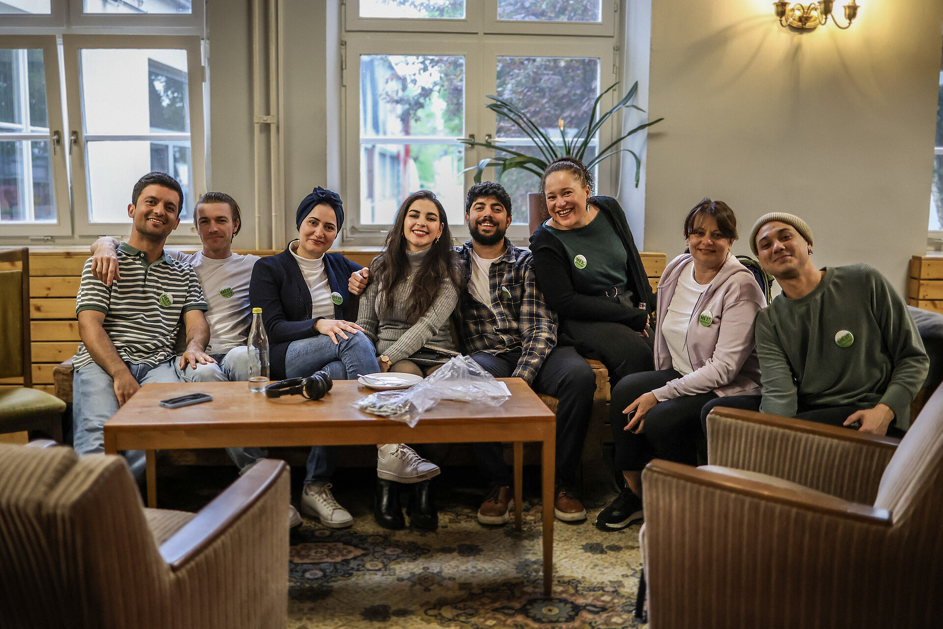 Projektschmiede_Teilnehmende des Projekts "MEETeinander" Gruppe von acht Personen sitzt auf einem Sofa und Stühlen in einem gemütlichen Raum mit Fenstern und einem Holztisch davor