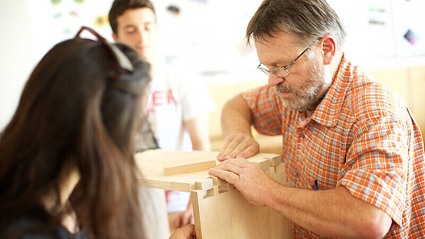 Mann in orange kariertem Hemd und zwei Personen arbeiten gemeinsam an einem Holzprojekt mit Holzkiste und Verbindungsstücken.
