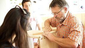 Mann in orange kariertem Hemd und zwei Personen arbeiten gemeinsam an einem Holzprojekt mit Holzkiste und Verbindungsstücken.