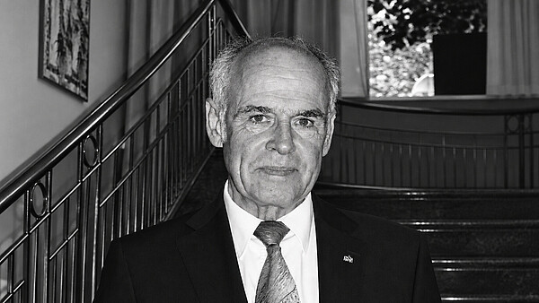 Schwarz-Weiß-Foto: Abgebildet ist ein älterer Mann in einem dunklen Anzug. Er steht vor einer Treppe.
