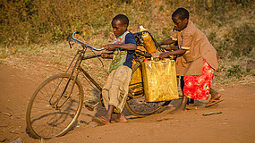Zwei Kinder mit Tüten auf einem Fahrrad im Sand