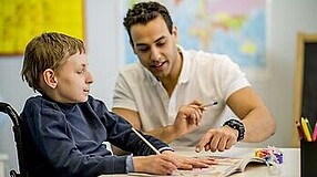 Junge im Rollstuhl sitzt mit Mann an einem Tisch und malt.