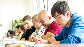 Menschen sitzen in einer Stuhlreihe am Tisch und schreiben.