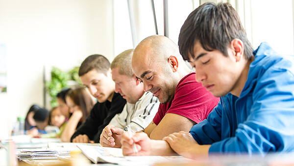 Menschen sitzen in einer Stuhlreihe am Tisch und schreiben.