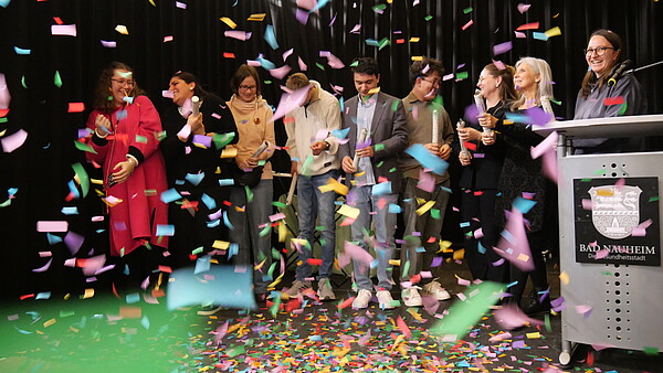 Auf der Bühne der Abschlussveranstaltung steht Tabea Trapp (3. von rechts) gemeinsam mit den Teilnehmenden des Programms abBi. Vor schwarzem Hintergrund fliegt buntes Konfetti.