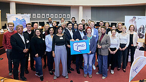 Gruppe Menschen stehen zusammen und halten ein Schild mit Aufschrift "JMD".