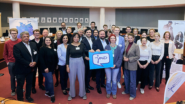 Gruppe Menschen stehen zusammen und halten ein Schild mit Aufschrift "JMD".