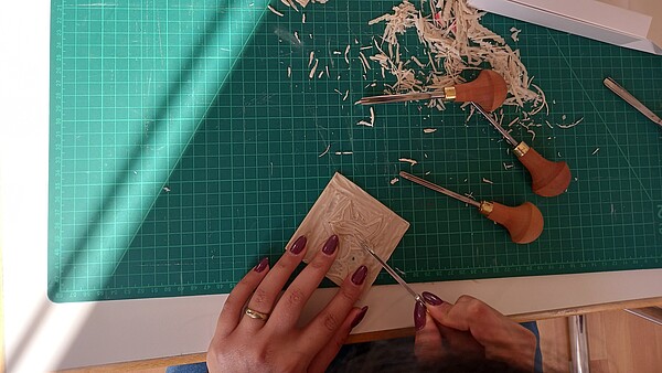 Zwei Hände bei der Arbeit: In der linken eine Platte, in der rechten ein Schneidewerkzeug. Auf der Arbeitsplatte sind Späne und weitere Werkzeuge zu sehen.