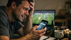 Ein Mann sitzt an einem Tisch und schaut auf ein Handy, das er in der Hand hält. Im Hintergrund sind ein unaufgeräumter Tisch und ein Fernsehgerät, in dem gerade ein Fußballspiel gezeigt wird.