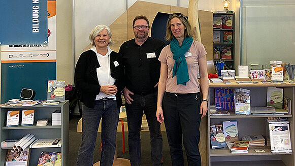 Drei Personen stehen vor einem Messestand. Neben ihnen und im Hintergrund befinden sich Regale mit Materialien wie Broschüren und Ähnliches.