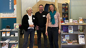 Drei Personen stehen vor einem Messestand. Neben ihnen und im Hintergrund befinden sich Regale mit Materialien wie Broschüren und Ähnliches.