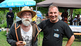 Zwei Männer stehen draußen auf einer Wiese. Der linke Mann trägt einen Sonnenhut und hält einen Keks in der Hand. Der rechte Mann trägt ein schwarzes T-Shirt mit der Aufschrift ‚IB‘. Im Hintergrund sind Sonnenschirme zu sehen
