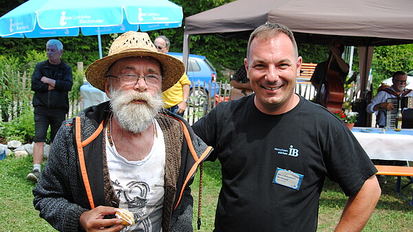Zwei Männer stehen draußen auf einer Wiese. Der linke Mann trägt einen Sonnenhut und hält einen Keks in der Hand. Der rechte Mann trägt ein schwarzes T-Shirt mit der Aufschrift ‚IB‘. Im Hintergrund sind Sonnenschirme zu sehen