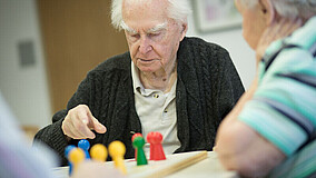 Ein älterer Mann sitzt zusammen mit anderen Personen, die nur teilweise zu sehen sind, an einem Tisch und spielt ein Brettspiel. Er trägt eine Strickjacke und wirkt nachdenklich.