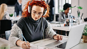 Person mit roten Locken und grauem Pullover sitzt am Schreibtisch, trägt weiße Kopfhörer und schreibt in ein Notizbuch neben einem Laptop.