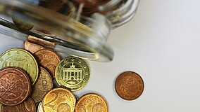 Zu sehen ist ein umgekipptes Glas mit Geldmünzen. Die Münzen liegen teilwiese auf einer weißen Fläche.