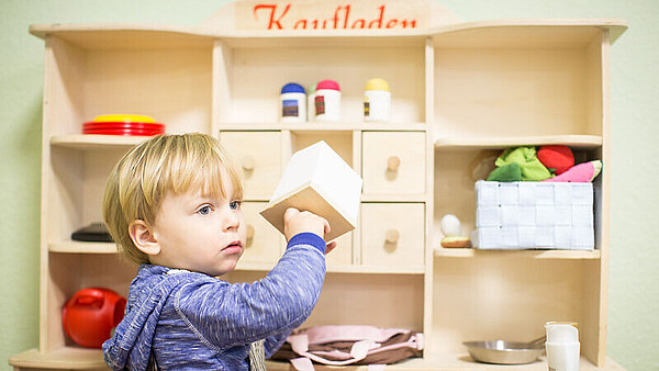 Kind steht vor einem Holzregal und hält einen weißen Würfel in der Hand