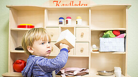 Kind steht vor einem Holzregal und hält einen weißen Würfel in der Hand