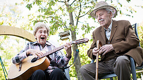 Ältere Frau und älterer Mann sitzen nebeneinander im Park. Sie spielt Gitarre, er lächelt.