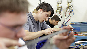 Mehrere Personen arbeiten an Werkbänken mit Werkzeugen, im Hintergrund sind Rohre und Ventile an der Wand zu sehen.