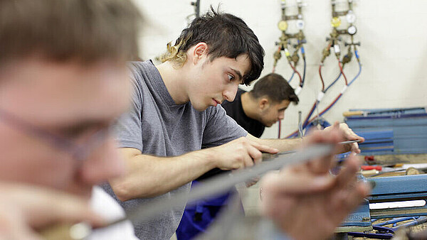 Mehrere Personen arbeiten an Werkbänken mit Werkzeugen, im Hintergrund sind Rohre und Ventile an der Wand zu sehen.